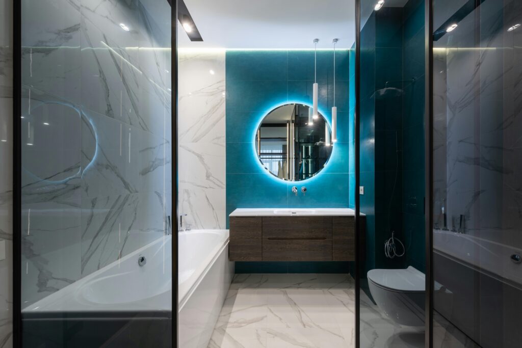 Round mirror with neon illumination hanging over sink in modern bathroom with white ceramic toilet bowl and bathtub